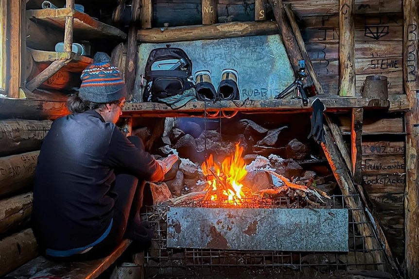 A bushwalker sits next to a fire in a hut