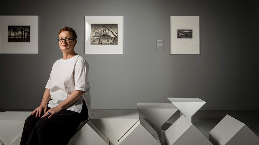 Woman with short cropped hair and glasses wearing why smock top and black pants in grey gallery with Escher prints on wall.