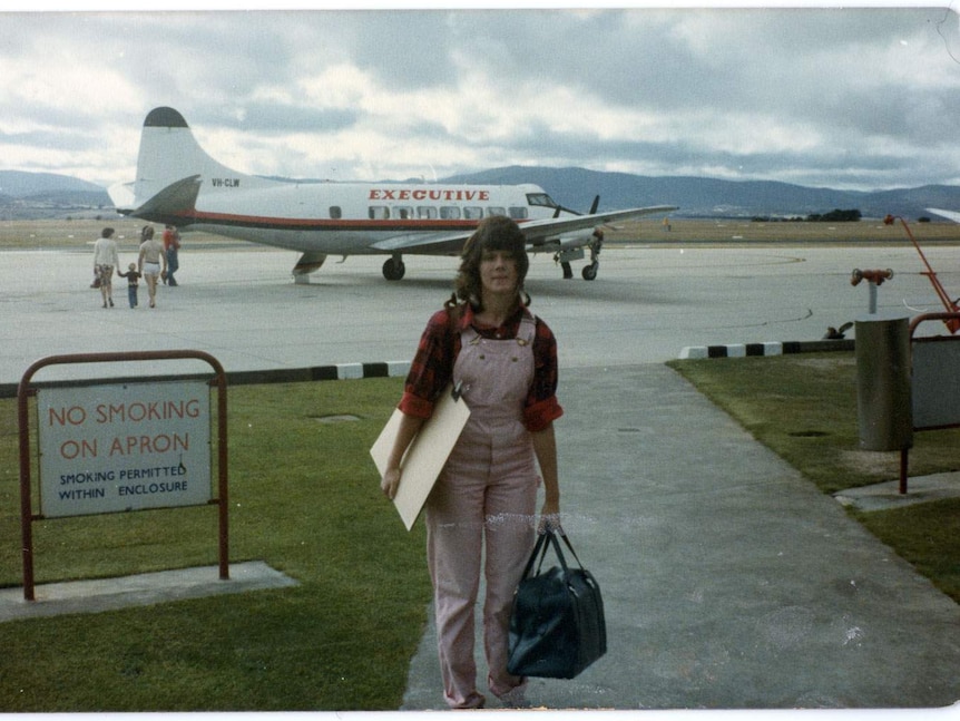 A woman faces the camera holding baggage. She is about to walk to an aeroplane waiting on the airstrip behind her.