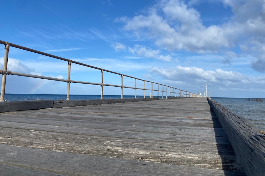 The boardwalk of the current Altona Pier is pictured with blue skies and clouds in the background.