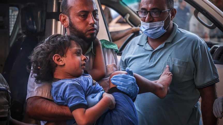 A young boy with tears on his cheeks is carried by a man out of a van