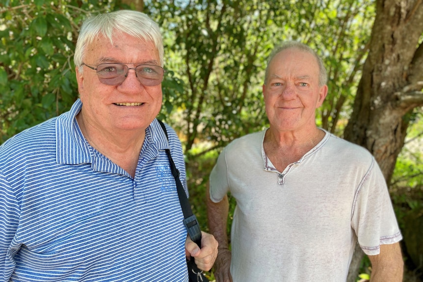 Two men in their 70s standing under a tree, smiling and looking at the camera
