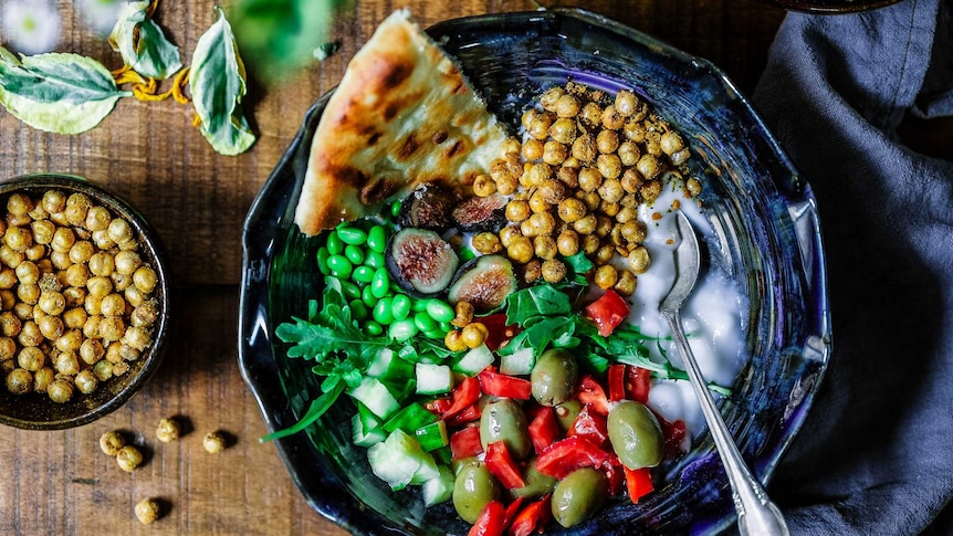A wooden table with bowls of chopped vegetables, chickpeas, bread and yoghurt.