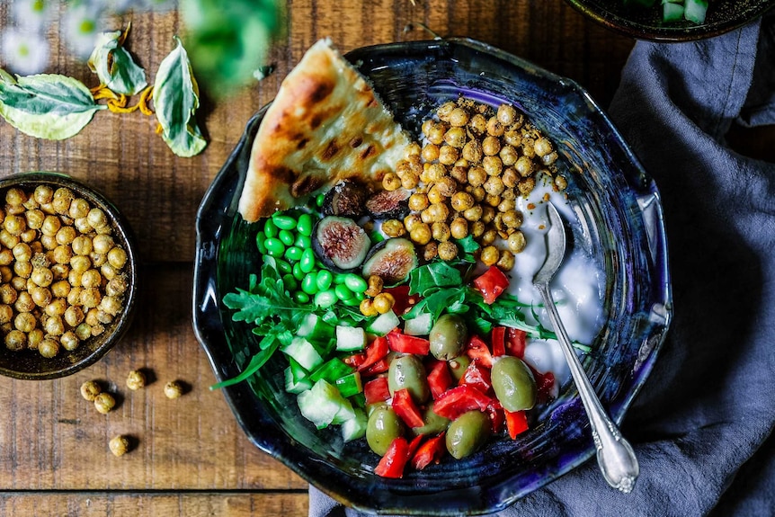 A wooden table with bowls of chopped vegetables, chickpeas, bread and yoghurt.