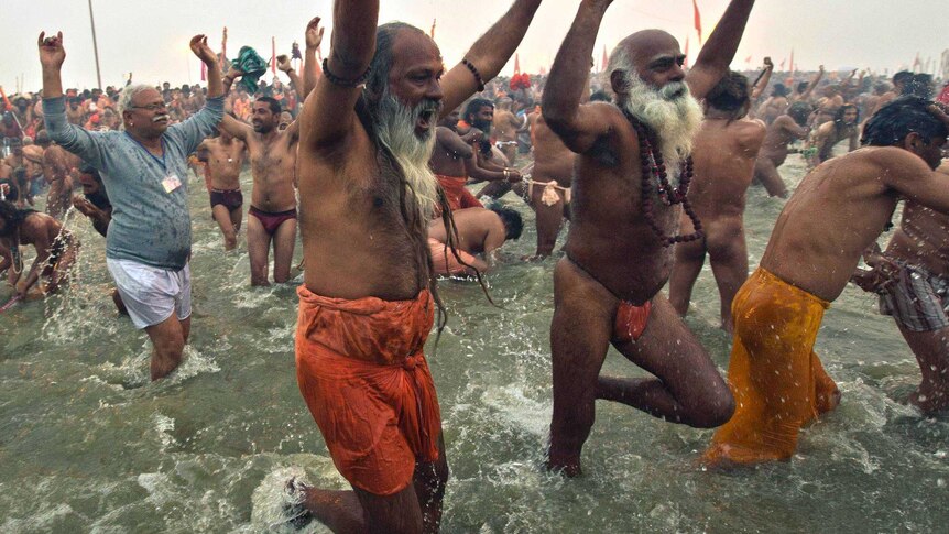 A large crowd of people, including sadhus, cheering as they walk into a holy river as part of the Kumbh Mela Festival