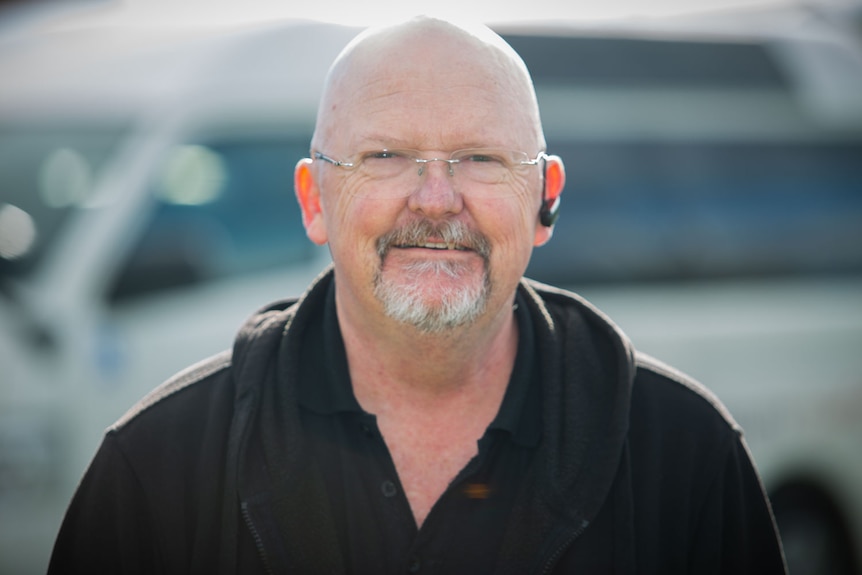 Bold portrait of a man with bald head and glasses, a white van blurred in background