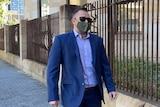 A man wearing a blue blazer, sunglasses and a face mask walks past an iron fence