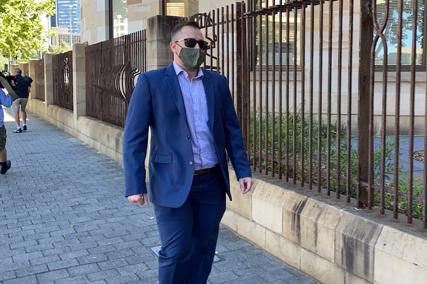 A man wearing a blue blazer, sunglasses and a face mask walks past an iron fence