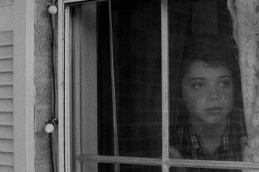 A black and white close-up still of Aidan Langford looking through a window in the 2018 film 1985.