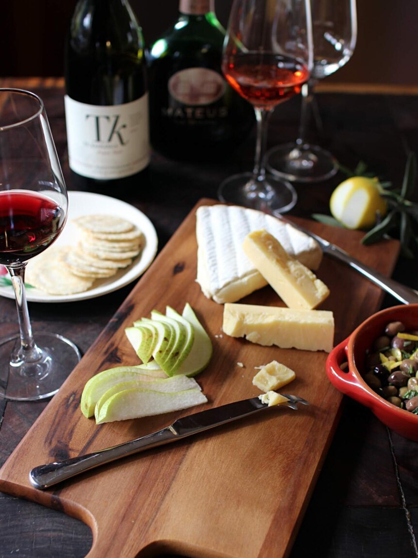 Two cheeses and sliced pear sit on a wooden board with red wine, olives and crackers nearby.