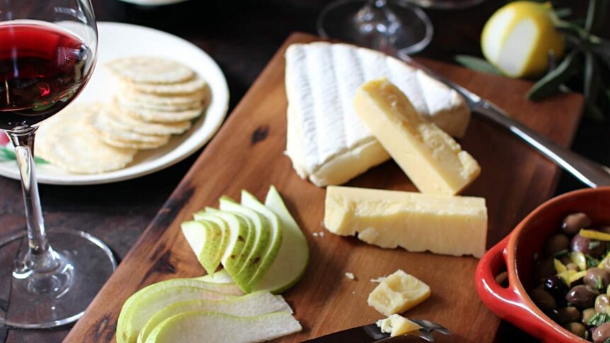 Two cheeses and sliced pear sit on a wooden board with red wine, olives and crackers nearby.