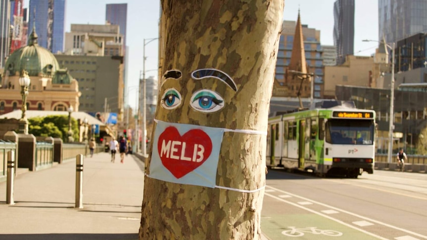 A sticker of eyes and eyebrows sit on a tree, with a mask with a love heart on it. In the background is a tram.