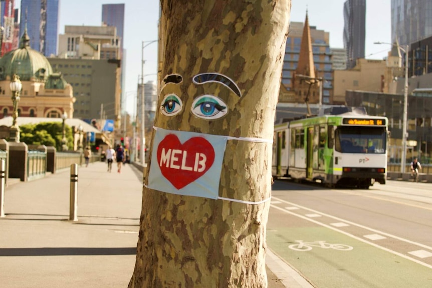 A sticker of eyes and eyebrows sit on a tree, with a mask with a love heart on it. In the background is a tram.