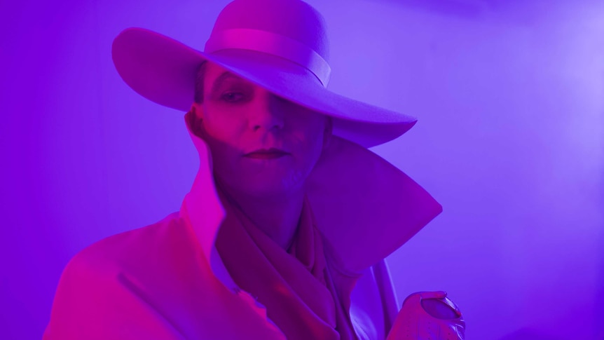The artist in heavy makeup under a wide brimmed hat and purple lighting, coat collar turned up.