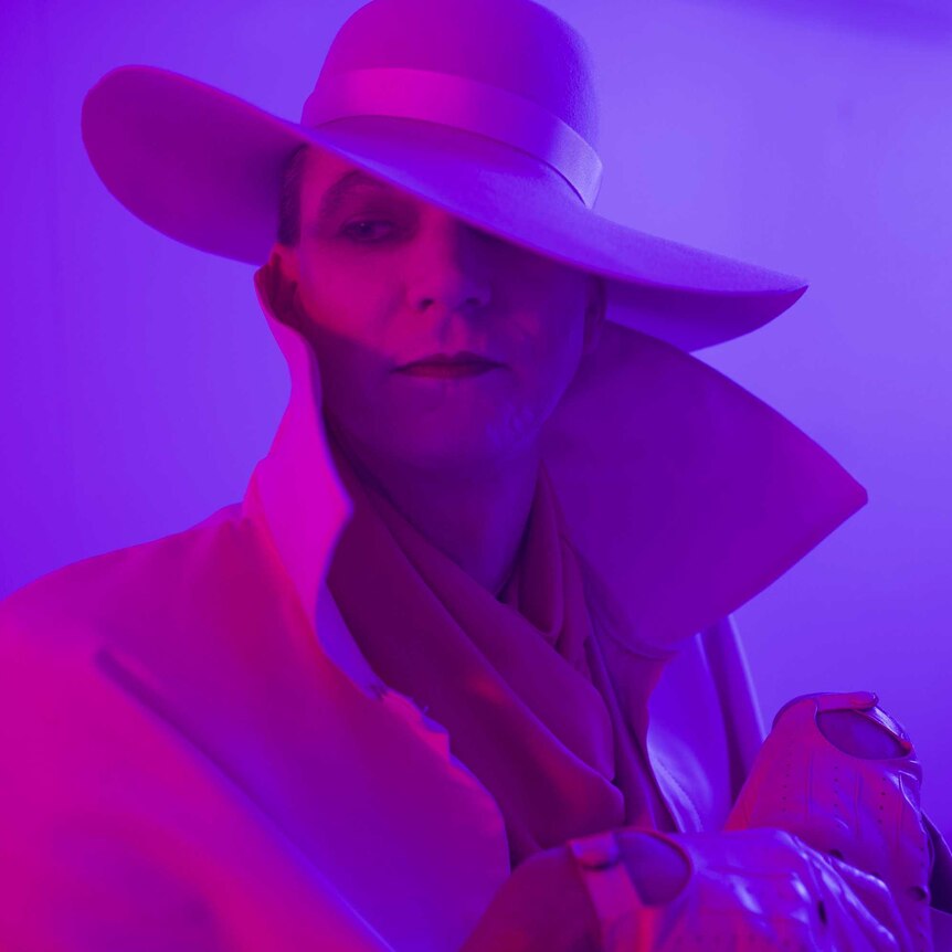 The artist in heavy makeup under a wide brimmed hat and purple lighting, coat collar turned up.
