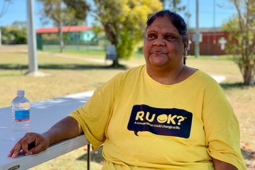 An Indigenous woman in a bright shirt with a dark logo on it sits down outside on a sunny day.
