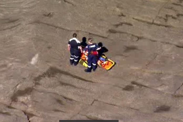 Two paramedics stand on a rock platform with a patient on a stretcher.