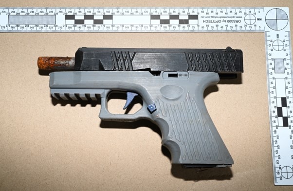 Un arma impresa en 3D de color gris y negro sobre una superficie plana junto a una regla cuadrada.