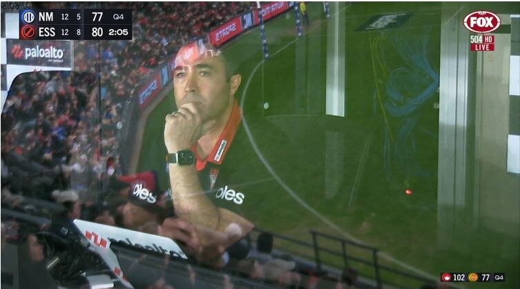 An image of a Essendon AFL coach pictured through the window of a coach's box during a game.