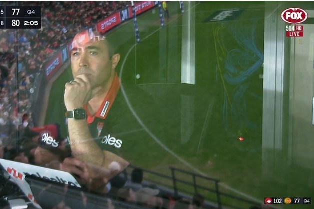 An image of a Essendon AFL coach pictured through the window of a coach's box during a game.