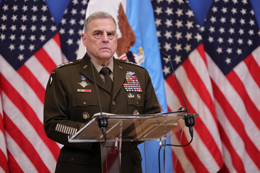 Mark Milley stands at a podium in military uniform with a row of American flags behind. 