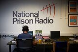 The back of a man in blue shirt wearing headphones, sitting at computer, with words National Prison Radio on wall behind him.