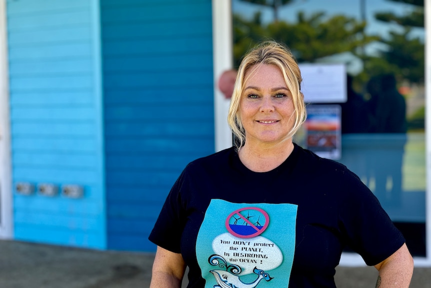 A smiling blonde woman wears a black t-shirt with a photo fo a fish, slogan and crossed out wind farms.