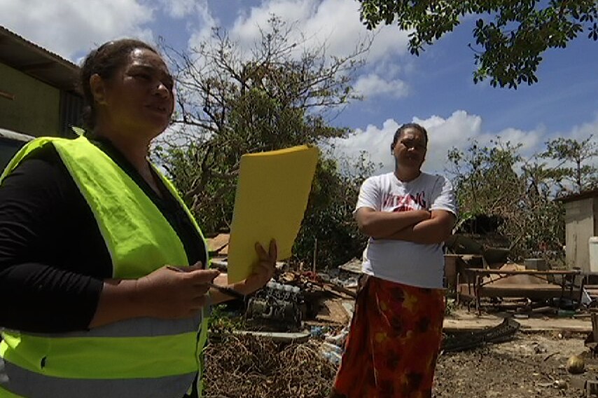 A person speaks to a Red Cross volunteer with damaged property in the background.
