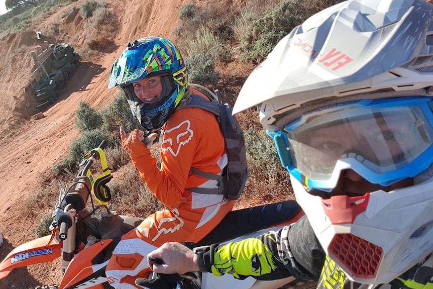 Girl in white helmet and goggles close up on right, with girl in orange overalls with helmet on motorbike in dirt scene 