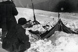 A black and white photo of a man crouching behind a wrecked tent in snow 