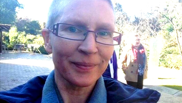 A woman with short hair and glasses looks at the camera, park in background.