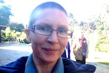 A woman with short hair and glasses looks at the camera, park in background.