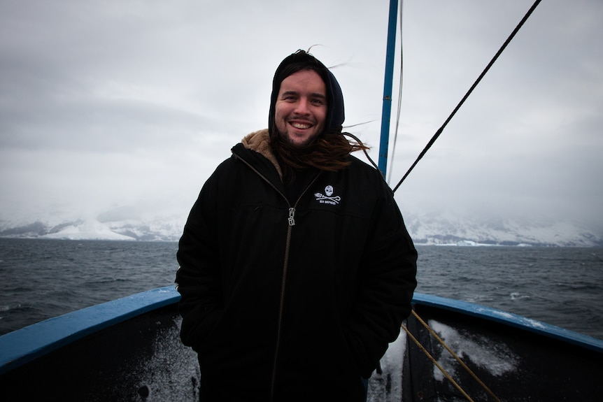  Alistair Allan wears a dark jacket with hood stands on the deck of a boat with Antarctica in the background.