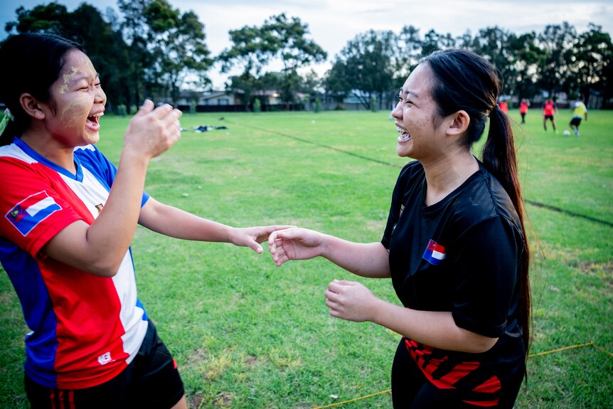 Two girls laugh and hold hands on a bright green footy oval. 