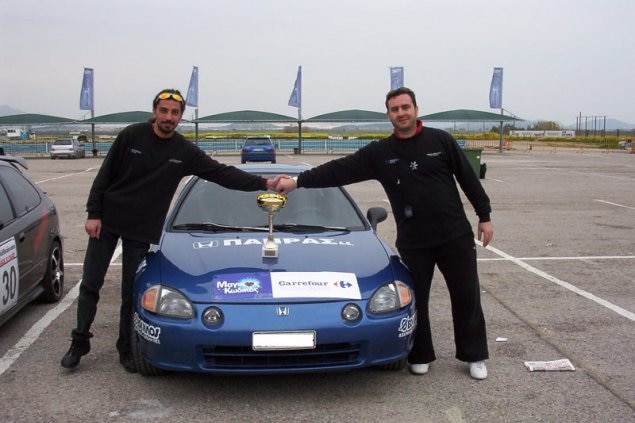 Fotis Larentzakis stands next to a racing car with a friend holding a gold trophy