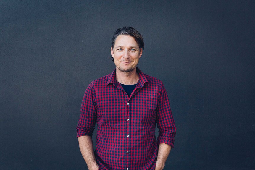 NORPA artistic director Julian Louis in a checkered shirt smiling and standing in front of a gray wall