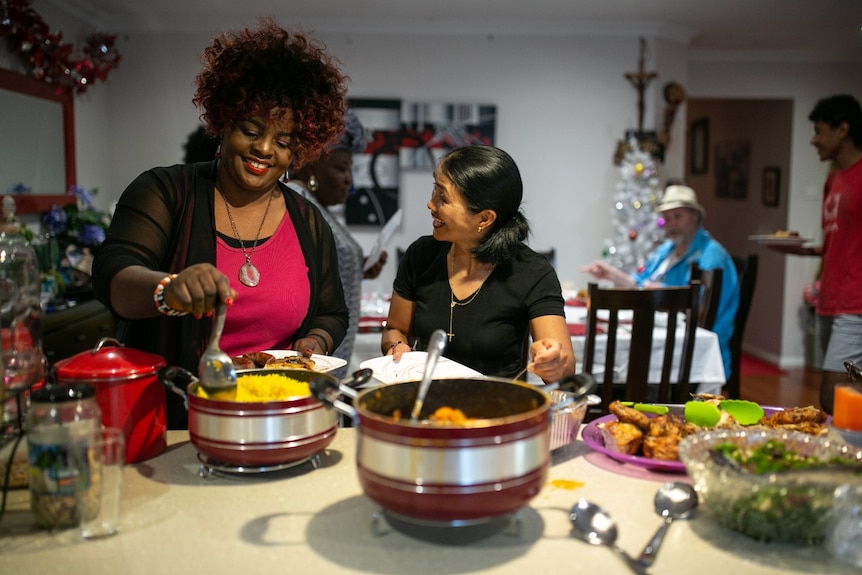 Two women smile as they serve themselves food from large serving dishes.