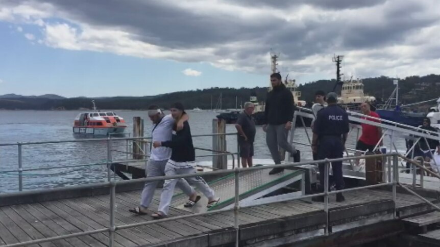 A man with a bandaged head leans on another man as they walk on a wharf