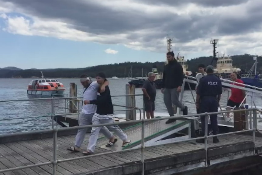 A man with a bandaged head leans on another man as they walk on a wharf