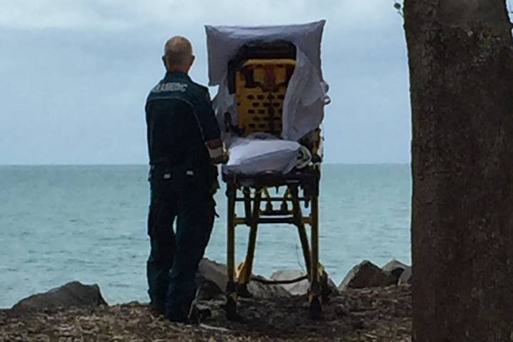 A Queensland Ambulance Service paramedic stands by a palliative patient in a stretcher by a beach in Hervey Bay