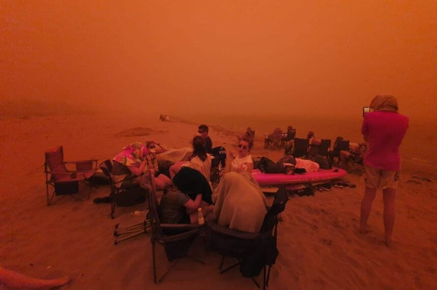 A group of people on a beach, the sky an intense orange from bushfire smoke.