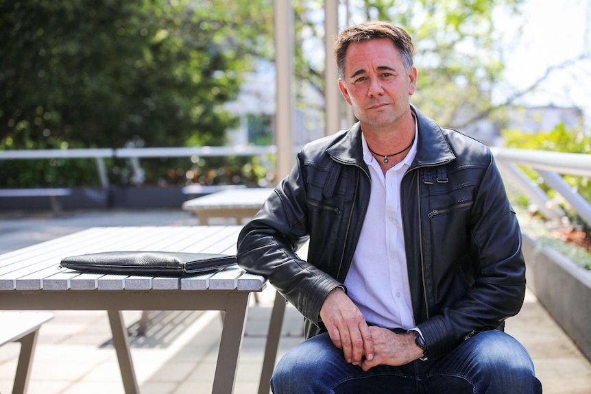 Jarrod Luscombe sits at a wooden bench and table.