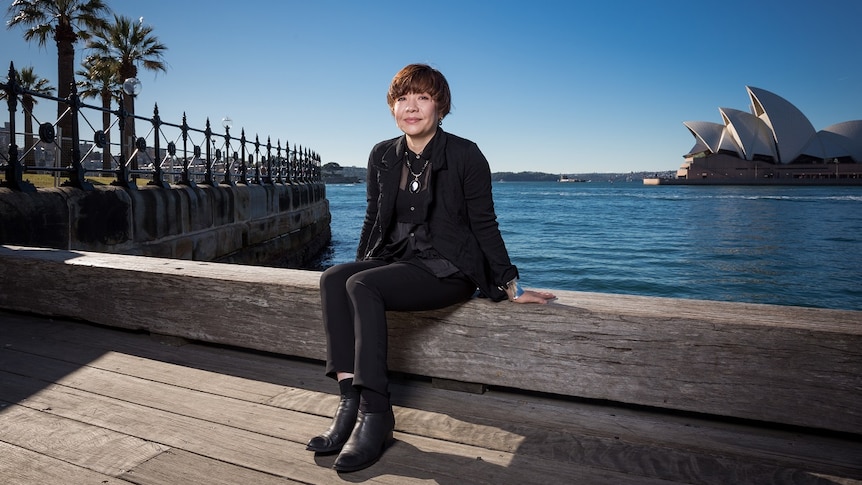 Biennale of Sydney director Mami Katoaka sits beside Sydney Harbour with Sydney Opera House in the distance behind her.