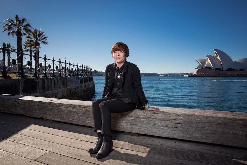 Biennale of Sydney director Mami Kataoka sits beside Sydney Harbour with Sydney Opera House in the distance behind her.