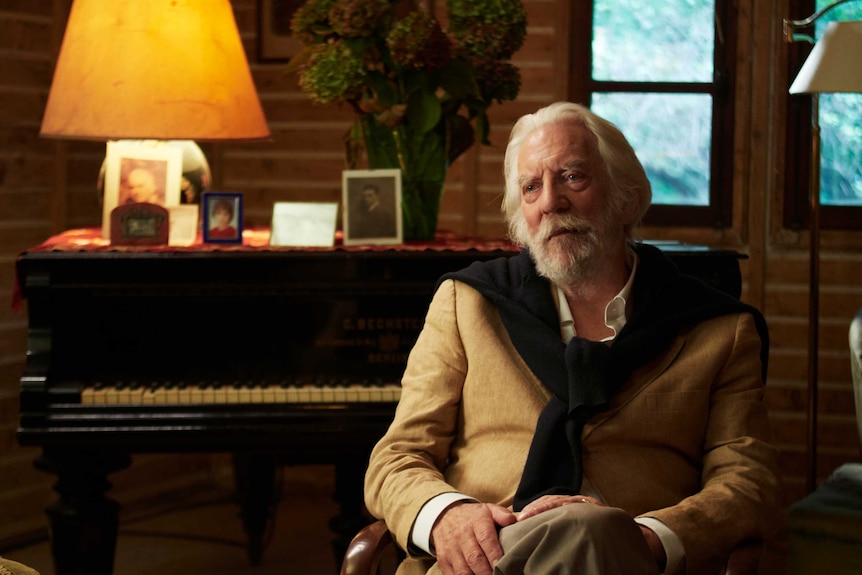 An older man with white beard and hair with jumper over shoulders of suits sits in cosy living room in front of grand piano.