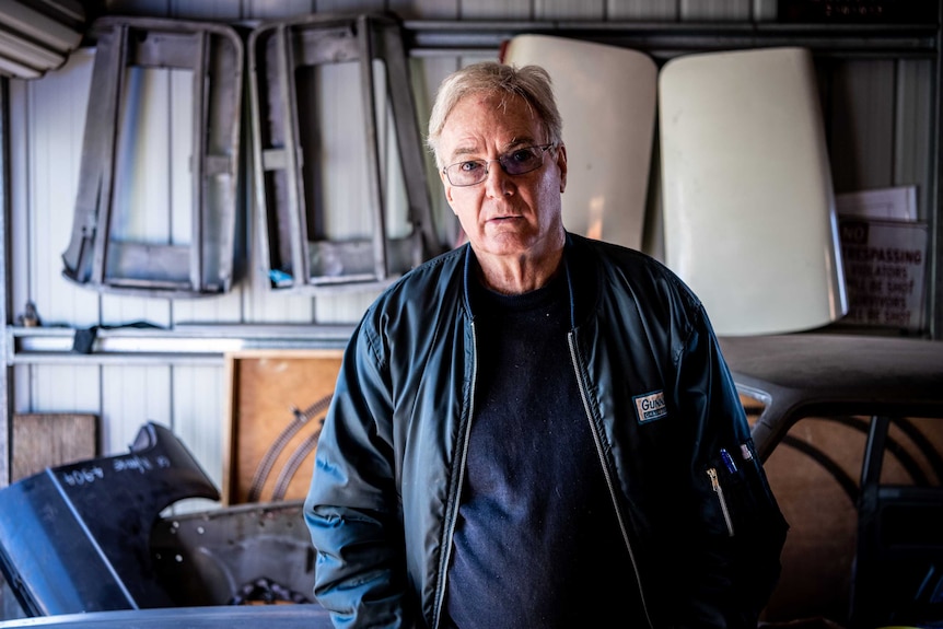 Greg Keenan looks into the camera while standing in garage with car panels behind him.