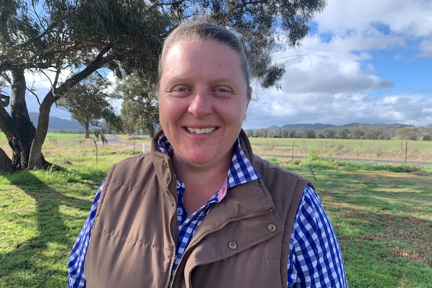 Kim Storey, wearing a brown vest over a blue and white checked shirt, smiles at the camera. Her property and a gum tree behind