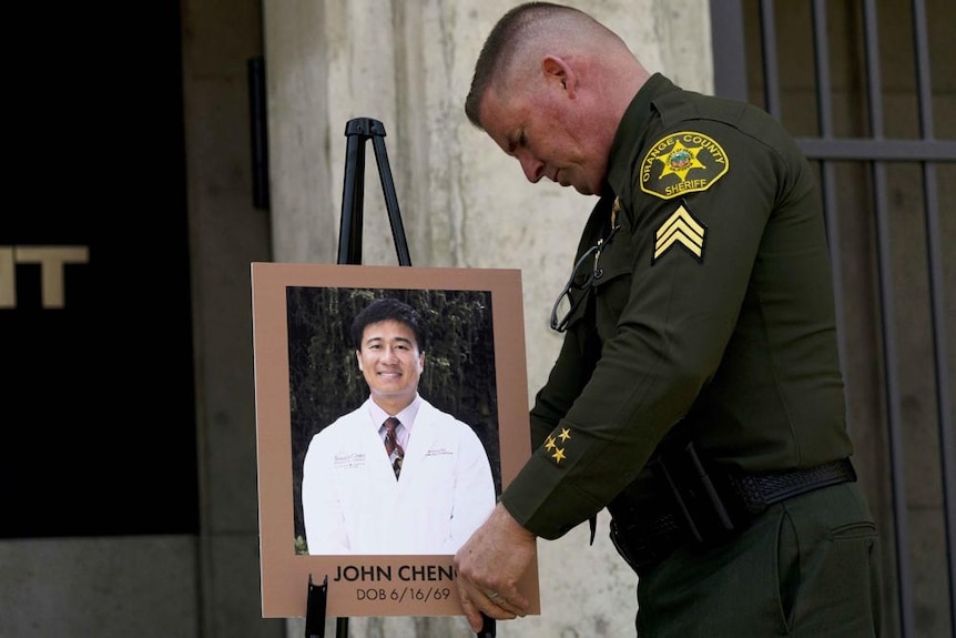 A Caucasian male police officer with a buzz haircut wearing an Orange County police uniform holds a photo of Asian man