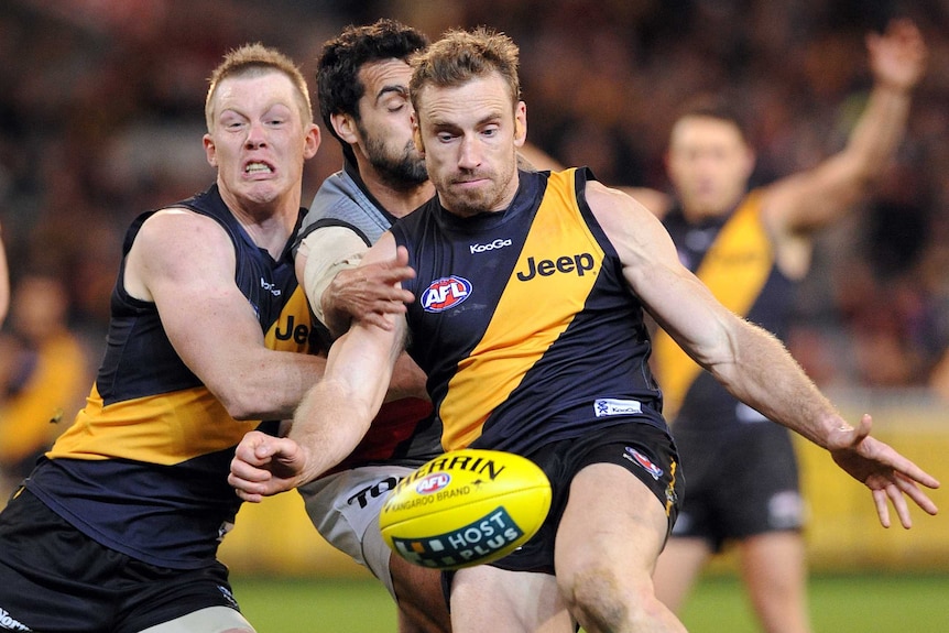 A Richmond AFL player tries to kick the ball with left foot with a Tigers teammate and Port Adelaide opponent next to him.