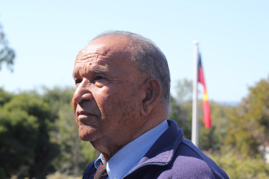 An indigenous man with an Aboriginal flag behind him, stares into distance.